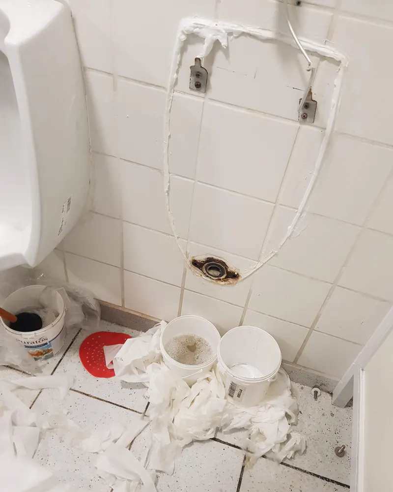 Muro de cerámica con el urinario retirado, dejando a la vista la conexión de la descarga y las cañerías de alimentación